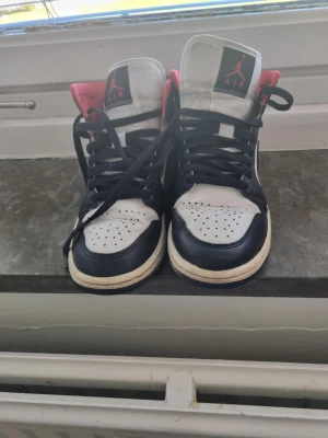 Air Jordan 1 svart/vit/röd - Snygga Air Jordan 1 Mid sneakers i vitt, svart och rött. Klassisk siluett med högt skaft, svart swoosh och röda detaljer på häl och plös. Ovandel i läder med perforerad tåbox och svart snörning. Perfekta för dig som gillar streetwear och ikoniska sneakers. I storlek 36,5 
