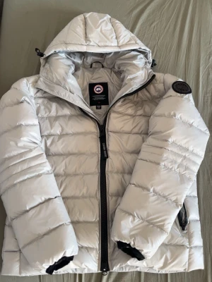 Vit dunjacka från Canada Goose - Canada Goose crofton - Snygg vit dunjacka från Canada Goose med huva och svart dragkedja. Jackan har quiltad design och klassisk logotyp på ärmen. Perfekt för våren och kalla dagar och har en modern, sportig look.                     Är storlek L men passar M också
