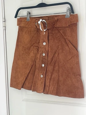 Brun mockakjol med bälte och knappar - Säljer en snygg brun kortkjol i mockaimitation med brett bälte och silverfärgat spänne. Kjolen har dekorativa knappar framtill och en lätt A-linjeformad siluett. Perfekt att styla med stickad tröja eller t-shirt.