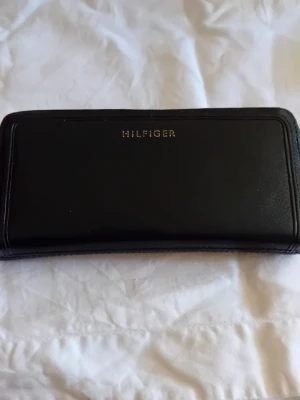 Svart plånbok från Hilfiger - Snygg svart plånbok från Hilfiger i stilren design med guldfärgad logotyp på framsidan. Plånboken är avlång och tillverkad i slätt material som ger en klassisk och tidlös känsla. Perfekt för dig som vill ha en enkel men elegant accessoar.Saknar tamp att dra med i myntfacket.
