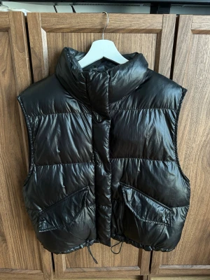 Svart dunväst med hög krage - Snygg svart dunväst med hög krage och två stora framfickor. Västen har en glansig finish, dragsko nedtill och breda quiltade paneler. Perfekt för lager på lager och ger en cool streetwear-look.