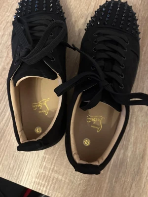 Svarta Louboutin sneakers med nitar - Säljer ett par svarta sneakers från Christian Louboutin med coola svarta nitar på tån och klassisk röd sula. Skorna har svarta snören och en lyxig känsla, perfekta för dig som vill sticka ut med din stil.