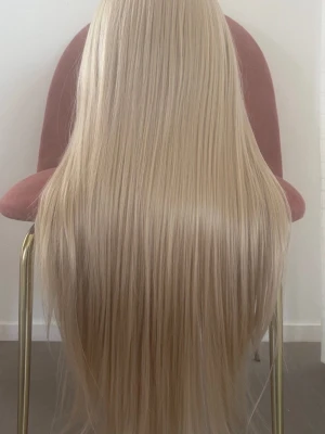 Lång blond lace front-peruk - En fantastisk blond peruk med långt, rakt hår som ger en elegant och naturlig look. Håret är gjort av mjuk och högkvalitativ syntetfiber som ser mycket realistiskt ut och känns lent. Perfekt för dig som vill ha ett fylligt och stilrent hår utan ansträngning.  Produktinformation: 	•	Färg: Blond       •     Storlek : Standardstorlek med      justerbara band  	•	Längd: 26 tum (ca 66 cm) 	•	Densitet: 180 % för en fyllig och naturlig volym 	•	Lace: 13 × 4 lace front med hög upplösning 	