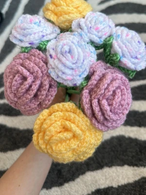 Virkat blomsterbukett i pastellfärger - Handgjord bukett med virkade blommor i lila, gult och vitt med färgglada prickar. Blommorna har gröna stjälkar bestående av pinnar med bomullsgarn lindat runt, även bladen är av samma garn. Själva roserna är gjorda i akrylgarn.