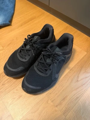 Svarta Nike sneakers mesh och syntet - Snygga svarta Nike sneakers med lågt skaft, tillverkade i mesh och syntetmaterial. Skorna har svarta snören och diskreta Nike-loggor på sidorna. Perfekta för en sportig och clean look.