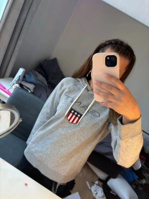 Grå hoodie från GANT med logga - Snygg grå hoodie från GANT med broderad logga och amerikanskt inspirerat märke på bröstet. Hoodien har dragsko i huvan och mjukt material som känns skönt mot huden. Perfekt för en avslappnad och trendig look.