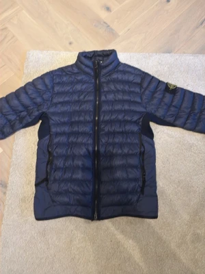 Mörkblå dunjacka från Stone Island - Snygg mörkblå dunjacka från Stone Island med ikonisk patch på ärmen. Jackan har två dragkedjeförsedda fickor. Nypris var strax över 5500kr. Har en lagning som syns på sista bilden. 