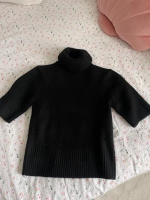 Svart stickad polotröja - Snygg svart stickad polotröja med halvlånga ärmar och bred ribbstickad kant nedtill och vid halsen. Perfekt för lager-på-lager och enkel att matcha med olika outfits. Mjuk och skön känsla.