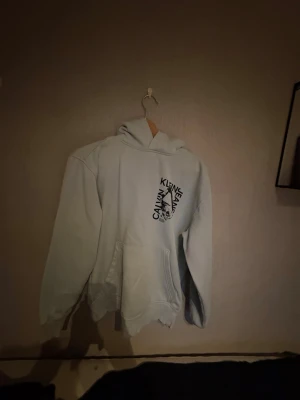 Ljusgrå hoodie från Calvin Klein - Snygg ljusgrå hoodie från Calvin Klein med svart logga tryckt snett över bröstet. Klassisk modell med huva och stor magficka framtill. Perfekt för en avslappnad och trendig stil. Mjuk och skön bomullsblandning.