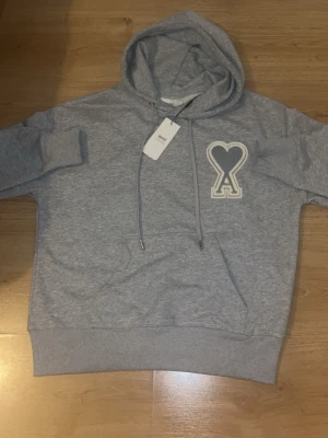 Grå hoodie från Ami Paris - Snygg grå hoodie från Ami Paris med stor broderad logga i form av ett hjärta och bokstaven A på bröstet. Klassisk känguruficka, dragsko i huvan och ribbade muddar. Perfekt för en avslappnad och trendig stil.