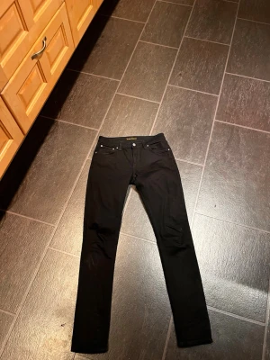Svarta slim fit jeans från Nudie - Svarta slim fit jeans från Nudie med klassisk femficksdesign och smal passform. Jeansen har svarta sömmar, knappgylf och läderpatch bak i midjan. Perfekta för en stilren och enkel look.