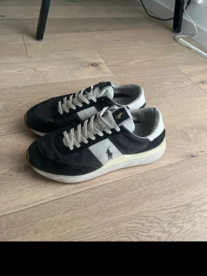 Svarta och vita sneakers från Polo Ralph Lauren - Snygga sneakers från Polo Ralph Lauren i svart och vitt med klassisk logga på sidan. Skorna har vita snören, låg profil och en bekväm sula. Materialmix av mocka och textil ger en sportig och stilren look.
