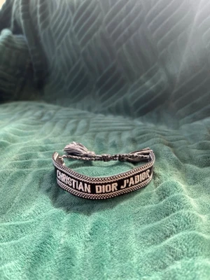 Christian Dior J'Adior armband - Snyggt vävt armband från Christian Dior med texten 'J'Adior' i vitt på svart bakgrund. Armbandet har justerbar knytning med tofsar i ändarna och är gjort i textilmaterial. Perfekt accessoar för att lyfta din outfit med en lyxig touch.