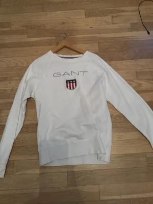 Vit sweatshirt från GANT med logga - Vit sweatshirt från GANT sitter som M-S typ med broderad logga och sköld på bröstet. Tröjan har rund halsringning och långa ärmar. Klassisk och stilren design med ribbade muddar och mjukt material. Perfekt för en avslappnad och snygg look.