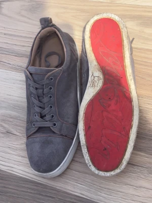 Christian Louboutins - Snygga gråa sneakers från Christian Louboutin med klassisk röd sula och diskreta sömmar. Skorna har lågt skaft, rund tå och är tillverkade i mocka med vita gummisulor. Perfekta för dig som vill sticka ut med exklusiva detaljer och ikonisk design.