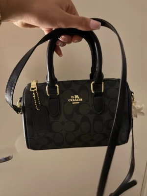 Svart handväska från Coach - Snygg svart handväska från Coach med klassiskt monogrammönster och guldfärgade detaljer. Väskan har både handtag och en lång axelrem, samt en dekorativ tag. Perfekt för dig som gillar stilrena accessoarer med lyxig känsla.