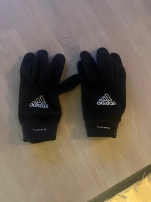 Svarta Adidas fotbollsvantar med grepp. - Svarta fotbollsvantar från Adidas med CLIMAWARM-tryck och logga på ovansidan. Fingertopparna har greppvänliga detaljer som funkar med touchskärm. Perfekt till fotbollsspelare.