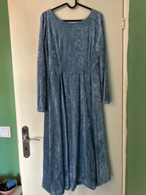 Blå långärmad sammetklänning - En långklänning i mjuk blå sammet med rund halsringning och långa ärmar. Klänningen har en enkel och elegant design med lätt utställd kjol, perfekt för dig som gillar en klassisk look med lite extra lyxig känsla.