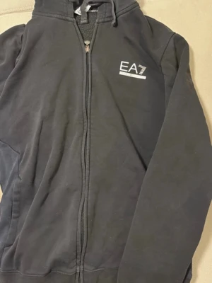 Svart hoodie från Emporio Armani EA7 - Snygg svart hoodie från Emporio Armani EA7 med dragkedja framtill och huva med dragsko. Logga på bröstet i vitt. Perfekt för en chill och stilren look. Tillverkad i mjuk bomull för maximal komfort.