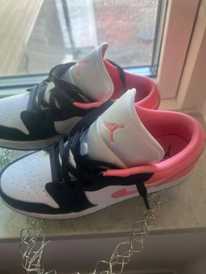 Air Jordan 1 Low svart/vit/rosa - Snygga Air Jordan 1 Low sneakers i svart, vitt och rosa. Skorna har klassisk låg siluett, svart snörning och rosa detaljer på swoosh, foder och Jumpman-logga. Ovandelen är i läder med perforerad tå för extra ventilation. Perfekta för dig som gillar streetwear och sportig stil.