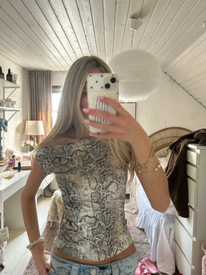 Offshoulder topp med ormmönster - Trendig offshoulder topp med grått och svart ormmönster. Sitter tight och har en croppad passform som framhäver midjan. Perfekt för dig som vill sticka ut med en edgy stil. Materialet känns mjukt och stretchigt.