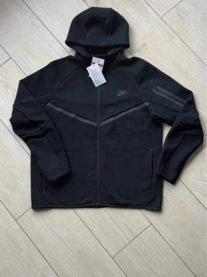 Svart Nike Tech Fleece hoodie - Nike Tech Fleece hoodie i svart med hel dragkedja och huva. Snygga detaljer med gråa linjer över bröstet och en extra ficka med dragkedja på ärmen. Tillverkad i mjukt och lätt material som håller värmen utan att bli tungt. Perfekt för en sportig och clean look.kommer utan lapparna