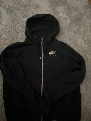 Svart Nike Air zip hoodie - Svart hoodie från Nike Air med dragkedja och huva. Framtill finns en liten Nike Air-logga i guld och på ryggen står det 'NIKE AIR' i stora bokstäver. Tillverkad i mjukt bomullsmaterial och har långa ärmar. Perfekt för en avslappnad och sportig stil.