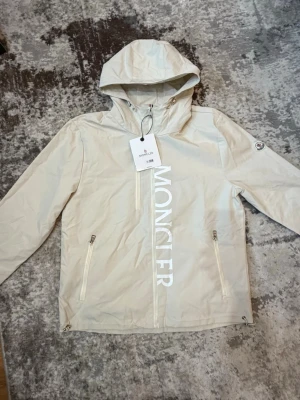 Beige vindjacka från Moncler - Stilren beige vindjacka från Moncler med huva och dragkedja framtill. Jackan har två fickor med dragkedja, stor vit Moncler-logga längs framsidan och en liten logotyp på ärmen. Lätt material, perfekt för blåsiga dagar.
