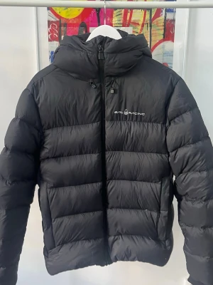 Svart dunjacka från Sail Racing - Snygg svart dunjacka från Sail Racing med huva och dragkedja framtill. Jackan har diskret logga på bröstet och är quiltad för extra värme. Perfekt för kalla vinterdagar och har en modern, sportig look. Storlek L men passar nog även större M. 