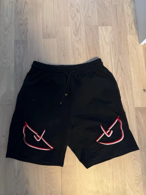 Fendi Shorts - Svarta shorts med elastisk midja och snörning. På båda benen finns broderade grafiska mönster i rött och vitt som ger en unik look. Perfekta för sommaren och avslappnade dagar.