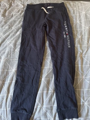 Svarta mjukisbyxor Tommy Hilfiger - Snygga svarta mjukisbyxor från Tommy Hilfiger med vit dragsko i midjan och tryckt logga längs ena benet. Byxorna har ribbade muddar vid bensluten och är gjorda i mjukt bomullsmaterial. Perfekta för chill dagar eller när du vill vara bekväm.