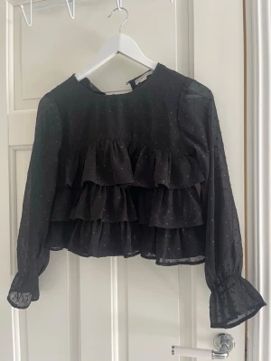 Svart volangblus från Gina Tricot - Svart blus från Gina Tricot med transparenta långärmar och små prickar i tyget. Blusen har flera volanger framtill och är tillverkad i ett lätt, luftigt material som ger en cool och trendig look.