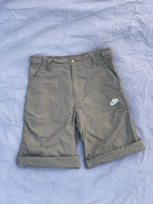 Kakhi shorts från Nike med logga - Snygga gröna shorts från Nike med vit broderad logga på ena benet. Klassisk passform med fickor på sidorna, bälteshällor och uppvikta benslut. Perfekta för varma dagar och avslappnad stil.