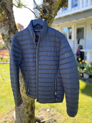 Blå vårjacka från Marc O'Polo - Snygg blå vårjacka från Marc O'Polo med quiltad design och två dragkedjefickor framtill. Jackan har hög krage och diskret logotyp på ärmen. Perfekt för kalla dagar och tillverkad i lätt, vadderat material.