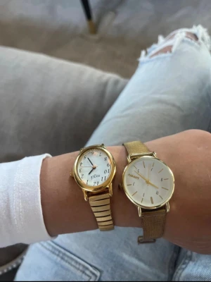 Två guldiga klockor!  - Två stilrena armbandsur i guld från Regal och Pilgrim. Båda har rund urtavla, en med klassiska siffror och länkarmband, den andra med minimalistiska markeringar och meshband. Perfekta accessoarer för att lyfta din outfit.