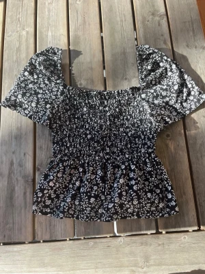 Svart blommig blus från H&M - Supersöt svart blus från H&M med vitt blommönster och puffiga korta ärmar. Smockad överdel som ger en snygg passform och lätt volang nertill. Använd ett fåtal gånger men har blivit för kort för mig är 170 cm.