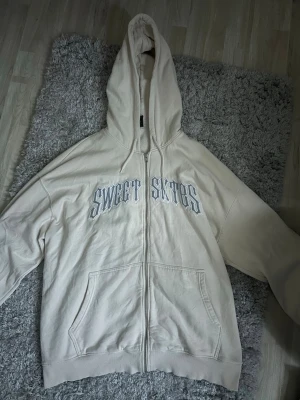 Beige hoodie från Sweet SKTBS - Beige zip-hoodie från Sweet SKTBS med trycket 'SWEET SKTBS' i vitt och grått på bröstet. Hoodien har huva med dragsko, två stora fickor fram och är tillverkad i mjuk bomullsblandning. Perfekt för en chill och avslappnad stil.
