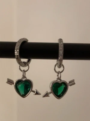Örhängen med gröna hjärtan och pil - Snygga örhängen i silverfärg med gnistrande gröna hjärtformade stenar och pil-detaljer. Ringarna har små klara stenar längs kanten för extra bling. Perfekt accessoar för dig som gillar unika och lekfulla smycken. Rostfri 
