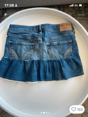 Blå jeanskjol med volang från Hollister - Säljer en blå jeanskjol från Hollister med volangkant nedtill och råa kanter. Kjolen har klassiska jeansfickor både fram och bak samt en liten logga på fickan. Perfekt för dig som gillar en avslappnad och trendig stil. Köpt på Vinted och förra ägaren har sytt den själv, bilderna är lite äldre och kjolen har en litet håll baktill🤗 