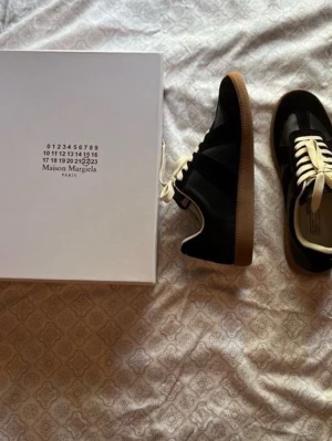 Maison Margiela svarta sneakers - Maison Margiela sneakers i svart läder och mocka med vita snören och klassisk gummisula. Snygg low-top modell med stilren design och diskreta detaljer. Perfekta för dig som gillar minimalistisk och exklusiv streetwear.