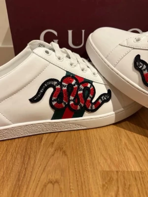 Gucci Ace vita sneakers med orm - Gucci Ace sneakers i vitt läder med broderad röd och svart orm på sidan och klassiska gröna och röda ränder. Snygg och ikonisk design med vita snören och präglad sula med Gucci-logga. Perfekta för dig som vill sticka ut med lyxig streetstyle.
