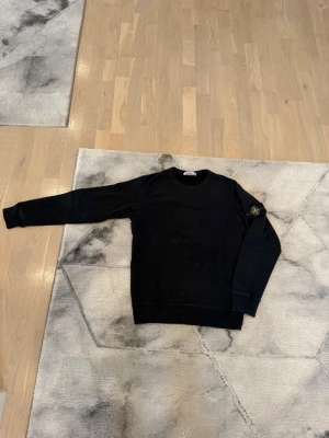 Svart sweatshirt från Stone Island - Säljer en svart sweatshirt från Stone Island med klassisk rund hals och ikonisk patch på vänster ärm. Tröjan har ribbade muddar vid ärmslut och nederkant, och är tillverkad i mjuk bomull för en skön och avslappnad look. Tröjan är lite blek men det är inget man tänker på