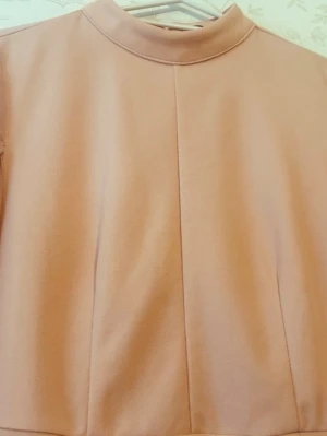 Lång ljusrosa klänning med capeärm - Elegant långklänning i ljusrosa med hög hals och dramatiska capeärmar i transparent tyg. Klänningen har en rak siluett och är tillverkad i ett mjukt, slätt material som ger ett exklusivt intryck. Perfekt för dig som vill sticka ut med en unik och stilren look.