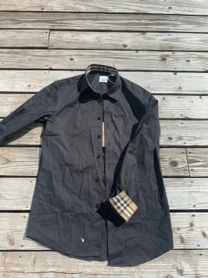 Svart skjorta från Burberry - Stilren svart skjorta från Burberry med klassiskt rutigt mönster på insidan av kragen och manschetterna. Skjortan har knappar framtill och broderad logga på bröstet. Perfekt för dig som gillar exklusiva detaljer och tidlös design.