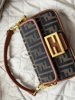Fendi Baguette monogram handväska - Säljer en ikonisk Fendi Baguette handväska i brunt och svart med klassiskt FF-monogram. Väskan har bruna läderdetaljer, avtagbar axelrem och en stor guldfärgad spänne framtill. Ej äkta 