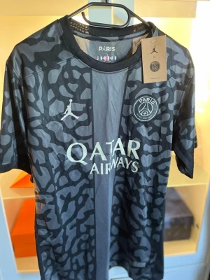 PSG x Jordan svart matchtröja - Snygg svart PSG matchtröja i samarbete med Jordan. Tröjan har ett unikt camomönster i grått och svart, rund halsringning och korta ärmar. Framtill finns PSG-logga, Jordan-logga och Qatar Airways-tryck. Perfekt för fotboll eller streetwear.