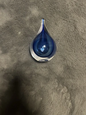 Blå glastdroppe prydnad - Säljer en dekorativ glastdroppe i klart och blått glas. Droppen har en elegant, avlång form och är perfekt som inredningsdetalj på hyllan eller skrivbordet. Färgen skiftar i olika nyanser av blått beroende på ljuset och ger ett stilrent intryck.