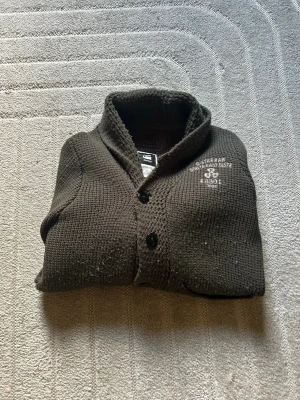 Mörkbrun kofta från G-Star RAW - Säljer en mörkbrun stickad kofta från G-Star RAW med två stora framfickor och tryckt logga på bröstet. Koftan har grovstickad struktur, sjalkrage och stängs med svarta knappar. Perfekt för lager på lager och chill stil.