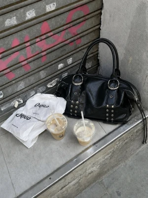 Svart handväska med nitar och spännen - Cool svart handväska i skinnimitation med silverfärgade nitar och stora spännen. Väskan har dubbla handtag och detaljerade sömmar, perfekt för en edgy streetstyle-look. Köpt secondhand! 