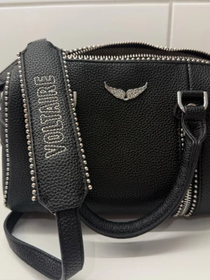 Svart Zadig & Voltaire handväska - Cool svart handväska i strukturerat läder från Zadig & Voltaire. Väskan har silverfärgade nitar längs kanterna, dubbla dragkedjor upptill och en bred axelrem med broderad logga. Dekorativt emblem med vingar framtill. Perfekt för dig som gillar edgy detaljer.